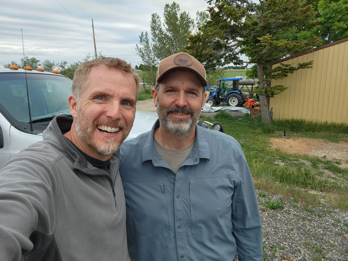 Jeremy and his friend Neal grinning in a farmyard selfie in Powell, Wyoming, tractor in the background