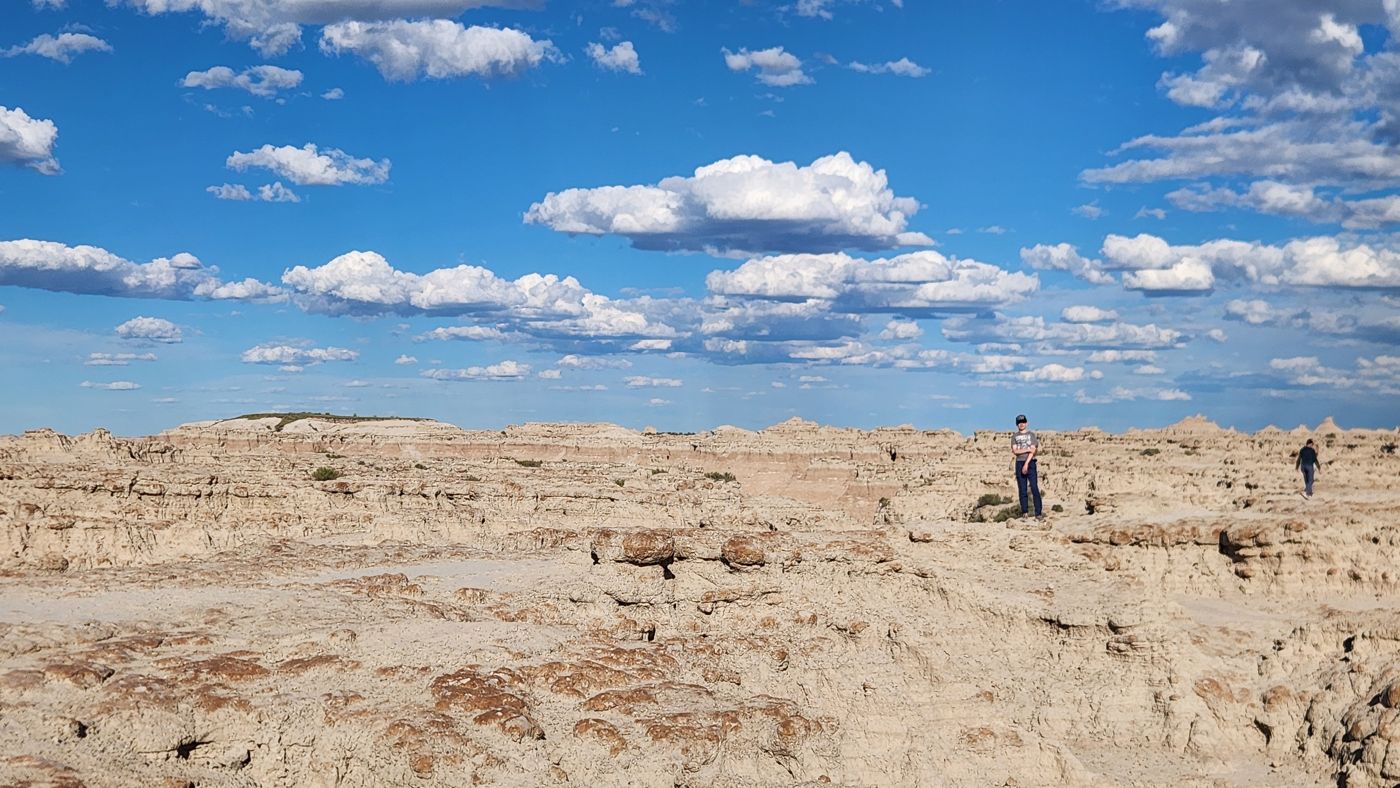 A wide Badlands vista with one boy standing small on the rim under a huge sky